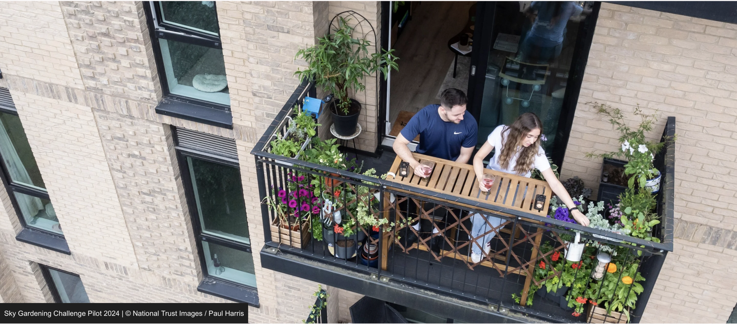 Aerial view of a couple sitting at a small wooden table on a narrow apartment balcony filled with potted flowers and plants, overlooking the side of a brick building.